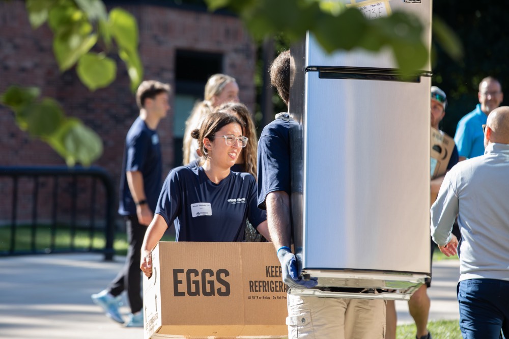 GVSU Alumnus carrying mini fridge with GVSU Alumna carrying a cardboard box labels "eggs"